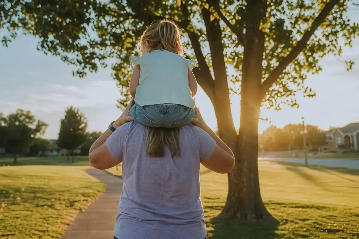 A woman carrying a small child on her shoulders along a park path at golden hour